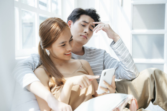 夫妻俩一起坐在窗户旁边的床上看智能手机里的照片。