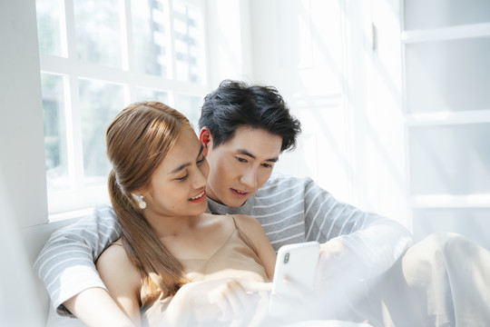夫妻俩一起坐在窗户旁边的床上看智能手机里的照片。