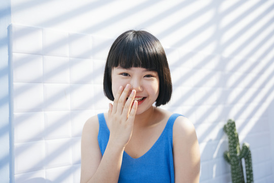 微笑美丽的鲍勃理发亚洲妇女在蓝色背心衬衫遮住她的嘴和笑白色瓷砖墙背景。