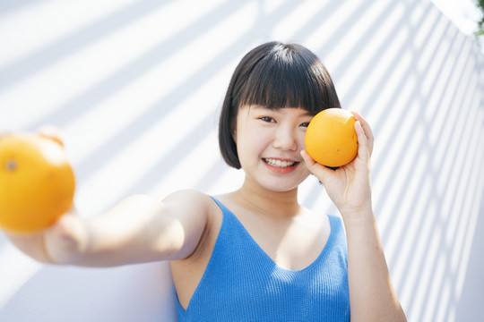 在白色背景下，穿着蓝色背心衬衫，手拿两个热带橘子和水果的美丽的鲍勃发型亚洲女人的特写肖像。健康的概念