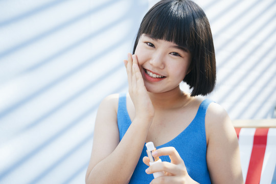 微笑美丽的鲍勃理发亚洲妇女在蓝色背心衬衫涂奶油乳液在她的脸上，坐在沙滩椅上白色背景。