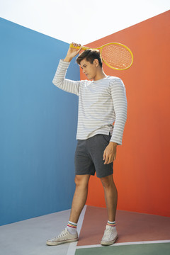 身着休闲服装、手持网球拍、背景为蓝橙色的迷人亚洲年轻男子。搞笑的姿势。打网球。站立姿势。