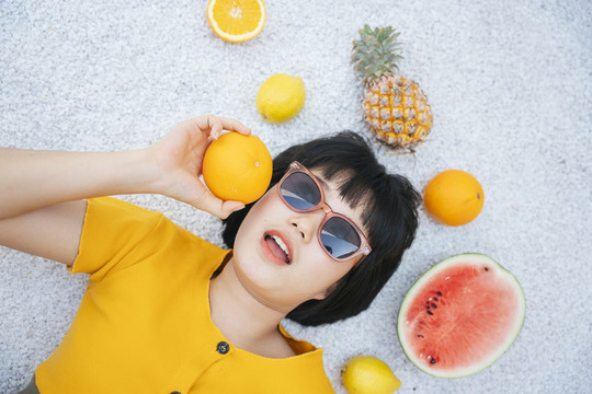 穿着黄色衬衫、戴着墨镜的亚洲年轻女子躺在地上，身上沾满了热带水果。
