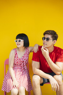 一对穿着红衬衫、戴着太阳镜的亚泰夫妇在橘黄色背景下坐在长凳上度过暑假。