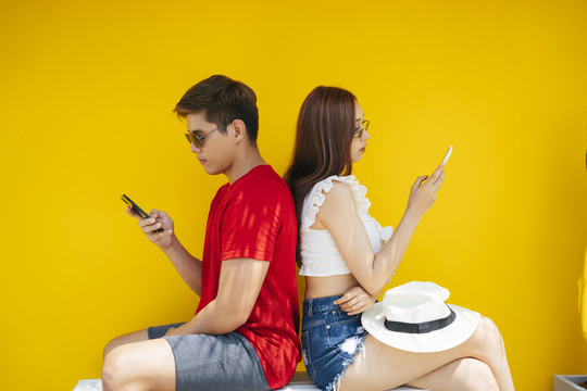这对夫妇背靠背地坐在一起，用智能手机，从不交谈。在橙色背景上。