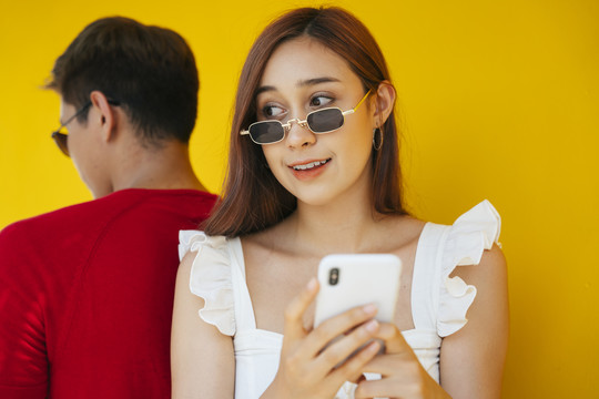 美丽的女人戴着眼镜用智能手机，坐在背靠背的年轻人红色衬衫在橙色的背景。
