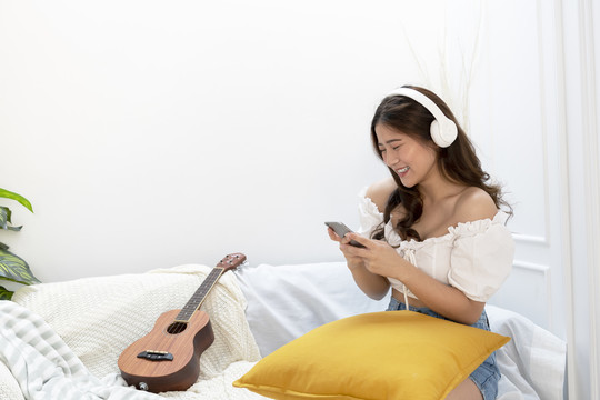 美丽的泰国亚裔年轻女子坐在客厅的沙发上，用智能手机的耳机听音乐。四弦琴躺在旁边。