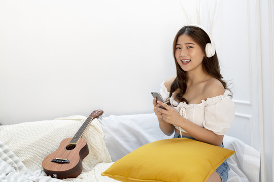 美丽的泰国亚裔年轻女子坐在客厅的沙发上，用智能手机的耳机听音乐。四弦琴躺在旁边。