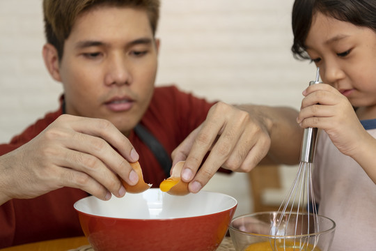 亚洲家庭烹饪活动-父亲将蛋黄从蛋壳中取出放入碗中，而儿子则在另一个碗中搅拌配料。
