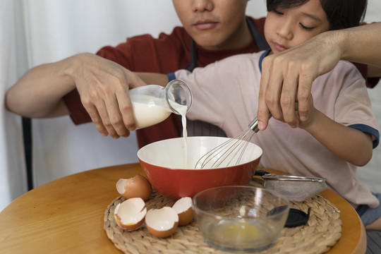亚洲家庭烹饪活动-父亲在厨房帮儿子做饭，将鲜奶倒入搅拌碗。