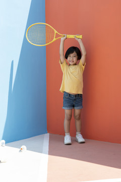 穿着黄色衬衫的可爱小男孩拿着网球拍，背景是五颜六色的蓝红色。地板上的羽毛球。把球拍举过头顶。