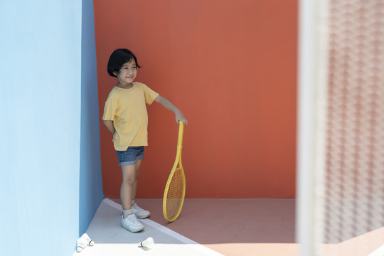 穿着黄色衬衫的可爱小男孩拿着网球拍，背景是五颜六色的蓝红色。地板上的羽毛球。拿着黄色网球拍。