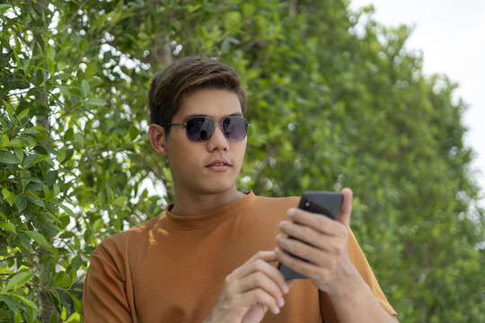 年轻英俊的亚泰男子戴着墨镜，在大自然中使用智能手机。