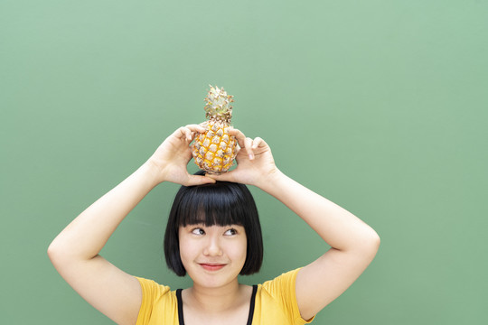 鲍勃发型泰国年轻女子举着菠萝在头上。隔离在绿色背景上。