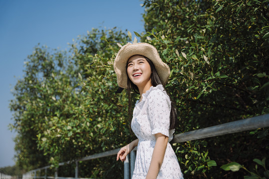 长发女子穿着连衣裙，戴着帽子，站在扶手旁，在蓝天下的树上微笑着。