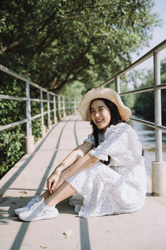 一个戴着帽子，穿着裙子的长发女人坐在小码头上，抱着她的膝盖。