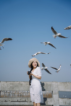 一位身穿白色连衣裙、头戴白帽的长发女子站在码头上，为蓝天上的海鸥拍照。