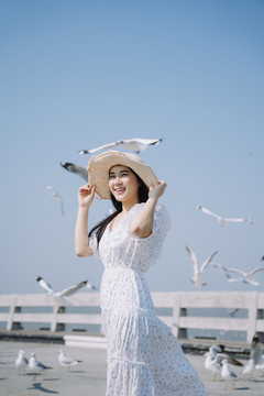 长发白衣白帽女子与海鸥一起站在蓝天下的阳光下。