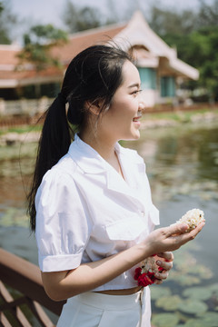 身穿白衬衫的黑马尾女人站在河边，手里拿着茉莉花环，河里有很多荷花。