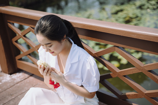 白衬衫马尾姑娘坐在河对岸的木桥上，手拿茉莉花环。
