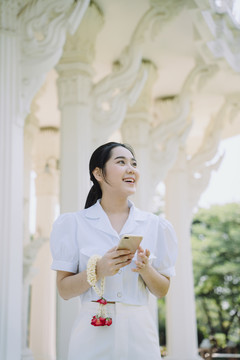 身穿白衬衫的黑马尾女孩把茉莉花环挂在手腕上，在庙里散步，用智能手机。