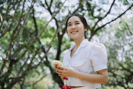 一个扎着马尾的黑女人，手持茉莉花环，穿过绿树园，走向寺庙。