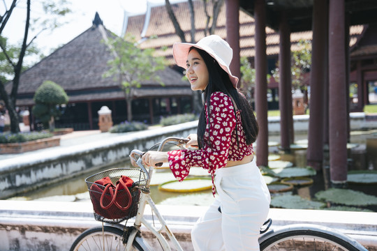 穿红衬衫戴红帽子的黑长发女孩骑着自行车来到传统的老房子里，用荷花在水池里，复制空间。