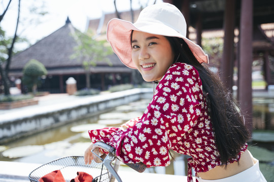 穿红衬衫戴红帽子的黑长发女孩骑着自行车来到传统的老房子里，用荷花在水池里，复制空间。