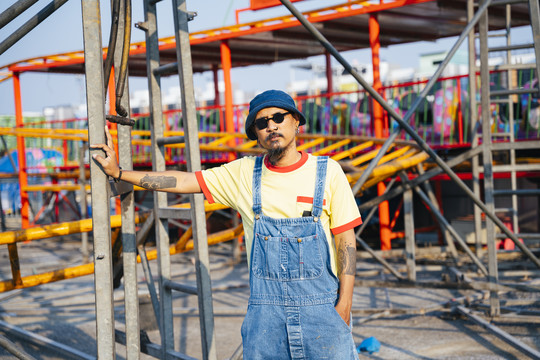 阳光明媚的日子里，一个穿着黄色t恤和牛仔工作服的男人站在废弃的游乐园里。