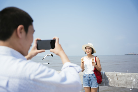 摄影师一名男子用智能手机拍下一名女子在海边拿着冰激凌。为blog和vlog制作内容。旅游理念。