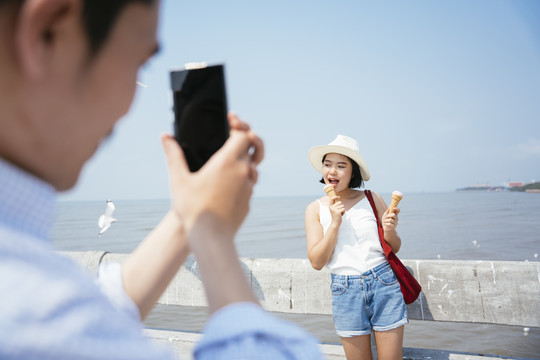 摄影师一名男子用智能手机拍下一名女子在海边拿着冰激凌。为blog和vlog制作内容。旅游理念。