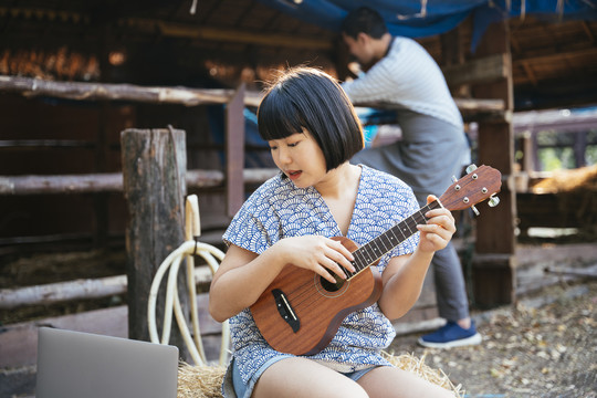 生态旅游-亚泰旅行者鲍勃发型女子在动物农场玩四弦琴吉他，看着笔记本电脑屏幕。乡下女孩。