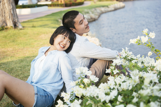 一对浪漫的亚泰夫妇背靠背地坐在河边公园的户外绿地上。前景是白色的花。