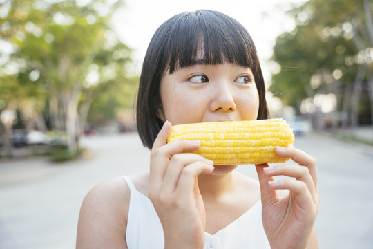 特写亚洲女人鲍勃发型吃煮玉米户外。健康的水果和蔬菜。