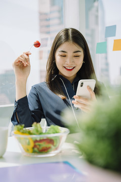 美丽的亚洲年轻泰国女子，深色长发，喜欢在办公室吃沙拉碗和使用智能手机。女商人喜欢蔬菜沙拉碗。午休时间