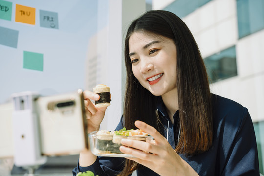 美丽的亚泰长黑发女士在办公桌旁用智能手机做健康食品录像机。城市背景。