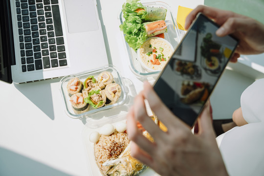 女性手持智能手机在办公桌上拍摄健康食品的俯视图。复制空间。