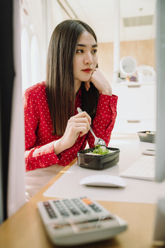 美丽的亚泰黑发红衫女子在家里电脑前吃健康食品。