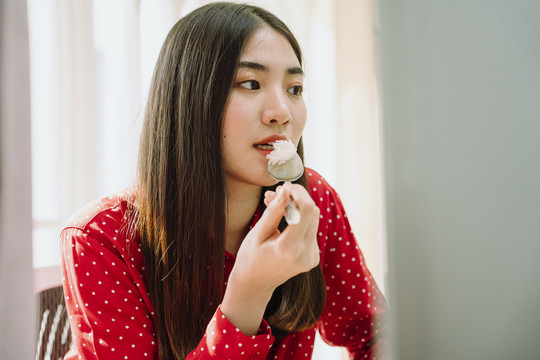 美丽的亚泰黑发红衫女子在家里电脑前吃健康食品。