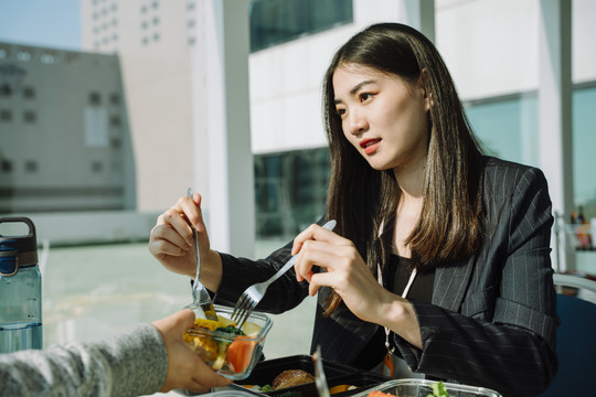 美丽的亚泰黑色长发女士，身穿黑色正式衬衫，在办公室与业务伙伴共进午餐。