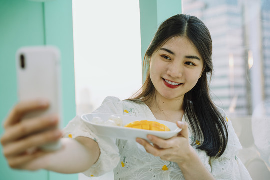快乐美丽的亚泰女人用智能手机自拍芒果糯米。