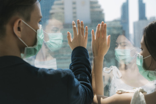 后视图-戴着医生面具的亚洲夫妇触摸玻璃窗，眺望城外摩天大楼。危机期间人们呆在家里。检疫。