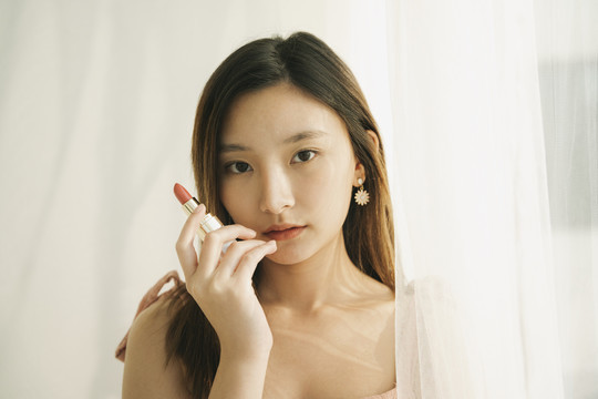美丽的泰国亚洲女人在白色房间里拿着口红化妆品。阳光和煦。