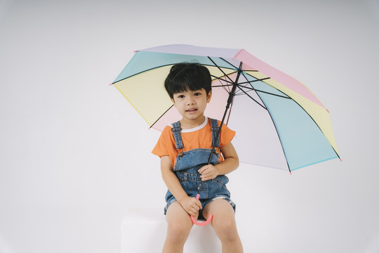 穿著牛仔褲工作服的可愛小男孩坐在工作室的白色街區(qū)上，手里拿著彩色粉彩傘。