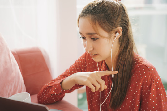 在家工作的概念-穿着红色睡衣衬衫的漂亮年轻女子用耳机和同事在笔记本电脑上交谈。检疫。
