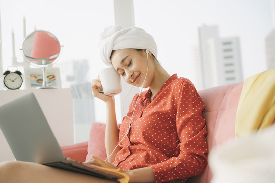 在家工作的概念-穿着红色睡衣衬衫，头上裹着毛巾的漂亮年轻女子坐在沙发上，拿着一杯咖啡，在笔记本电脑上