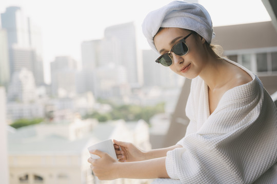 复制空间。侧视图-在公寓阳台，穿着白色浴袍的年轻女子拿着一杯咖啡。