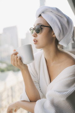 特写肖像——身穿白色浴袍、戴着墨镜的年轻女子在城市背景上拿着一杯咖啡喝酒。