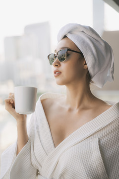 特写肖像——身穿白色浴袍、戴着墨镜的年轻女子在城市背景上拿着一杯咖啡喝酒。