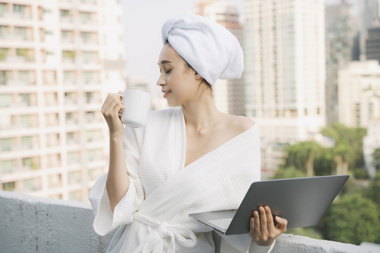 穿着白色浴袍的年轻女子早上在家拿着笔记本电脑喝热咖啡。在家工作。